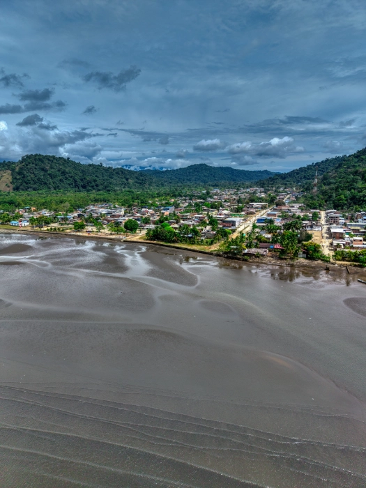 Vista aérea de un pueblo costero asentado entre colinas selváticas, con una extensa playa de arena oscura durante la marea baja y canales de agua que forman patrones en la superficie.