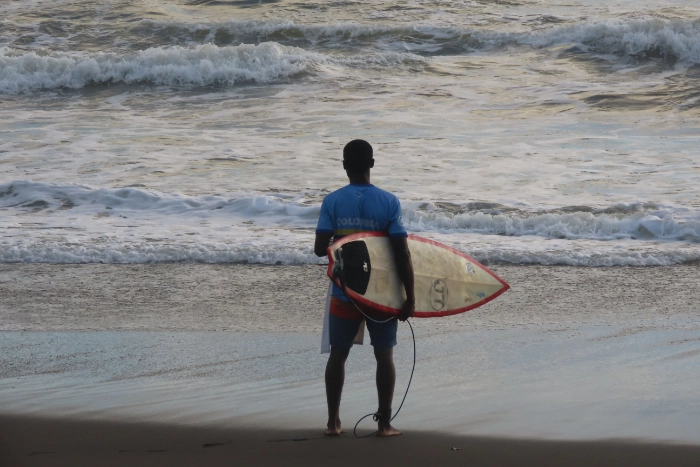 Surfista de espaldas con camiseta de Colombia sosteniendo su tabla de surf en una playa de arena oscura, contemplando las olas del mar pacífico.