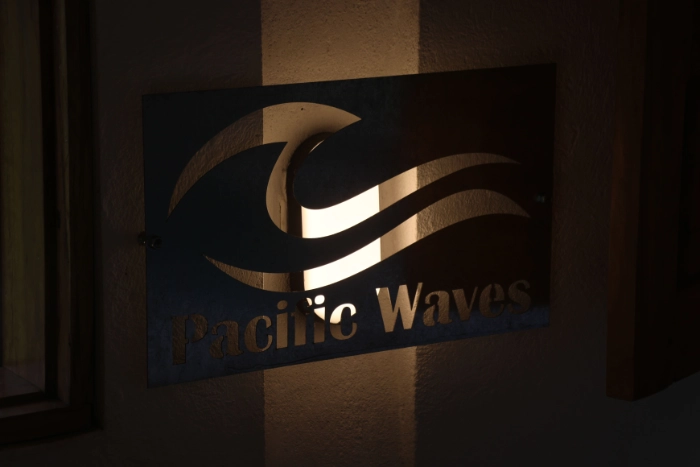 Letrero metálico retroiluminado de Pacific Waves con un diseño calado en forma de olas del mar, montado en una pared exterior durante la noche.