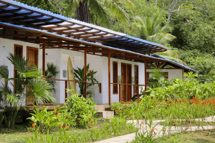 Fachada de las cabañas blancas de Pacific Waves con pasillos de madera, vegetación tropical exuberante al frente y una tabla de surf apoyada en la pared.