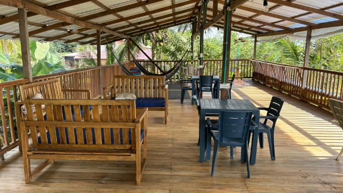 Área social abierta y techada con pisos de madera, equipada con sillones de madera, mesas con sillas plásticas y hamacas al fondo para el descanso.