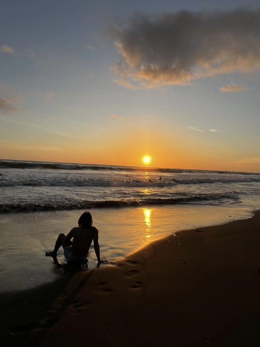 Silueta de una persona sentada en la orilla de una playa de arena oscura durante el atardecer, con el sol reflejándose en el agua del mar.