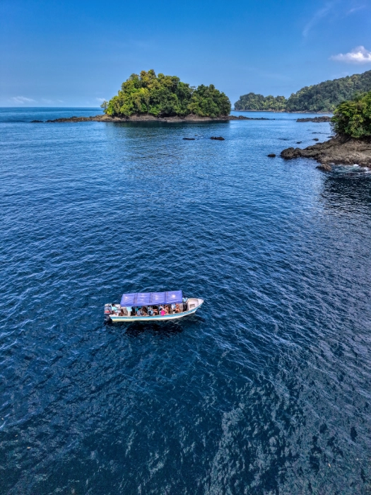 Vista aérea de una lancha de turismo con techo azul navegando sobre aguas profundas y azules del océano, cerca de un pequeño islote cubierto de árboles verdes.