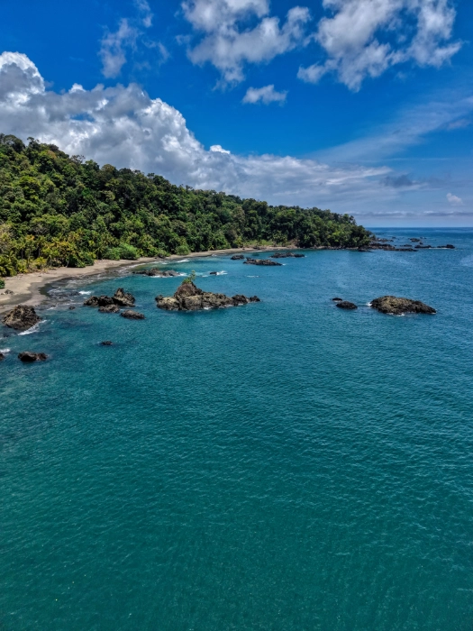 Panorámica aérea de una bahía con agua de color azul turquesa brillante, una costa selvática y varias formaciones de rocas oscuras que sobresalen de la superficie del mar bajo un cielo azul con nubes blancas.