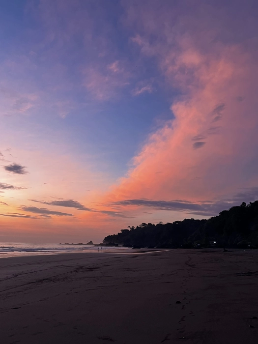 Atardecer en una playa tropical con el cielo teñido de tonos morados, azules y una gran franja de nubes rosadas. Al fondo se observa una costa cubierta de densa vegetación y la silueta de dos personas caminando cerca de la orilla del mar.