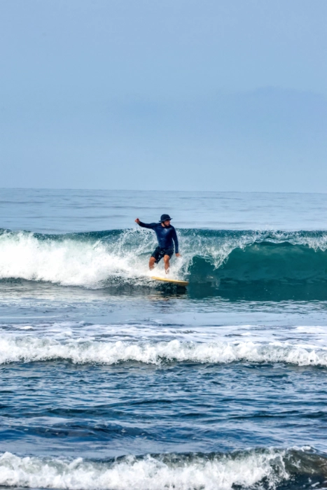 Un surfista con licra oscura y gorro manteniendo el equilibrio sobre una tabla de surf amarilla mientras desciende por la cresta de una ola en el océano.