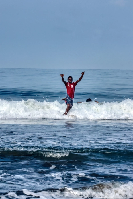 Un surfista celebrando sobre una ola en el mar, vistiendo una camiseta roja y pantaloneta azul con estampados, con los brazos en alto haciendo el gesto de 'shaka' bajo un cielo despejado.